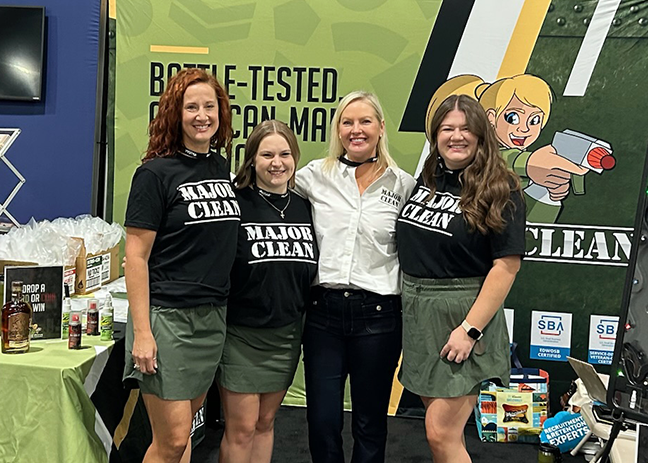 Four people stand side by side at a trade show booth, posing in front of a green backdrop that reads “Battle-Tested Performance.” Three individuals wear black “MAJOR CLEAN” T-shirts with green skirts, while the person in the center wears a white button-down shirt. A table with branded products and signage is visible to the left, and promotional materials and boxes appear on the right, reinforcing the product-focused display.