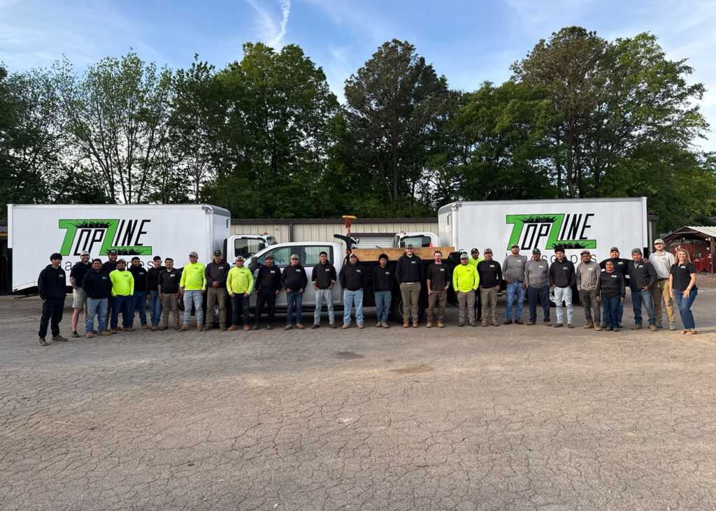 Large group of employees standing side by side in a parking lot in front of two white trucks and a pickup, each with the “TopLine” logo, with trees and a blue sky in the background.