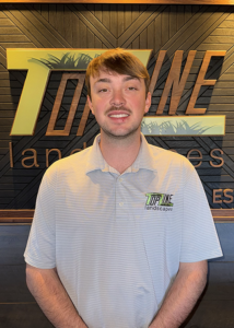 Individual standing indoors in front of a wall with a large “TopLine Landscapes” logo, wearing a light-colored polo shirt with the company logo embroidered on the chest.