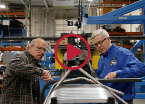 Two people work together at an automated tube-bending machine inside a manufacturing facility. One person in a blue company shirt and another in a plaid shirt examine curved metal tubing positioned on the machine. The industrial setting includes blue and yellow equipment, metal components, and overhead signage reading “Welding.”