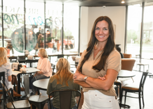 Janna Tucker stands with arms crossed in a bright restaurant; diners are seated at the tables behind her.