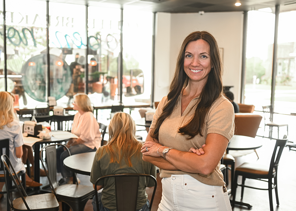 Janna Tucker stands with arms crossed in a bright restaurant; diners are seated at the tables behind her.