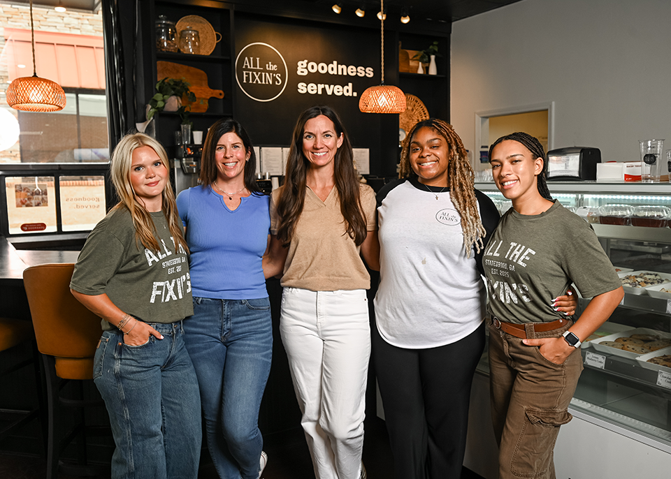 Janna Tucker poses with four staff members in front of a café counter with “All the Fixin’s” sign and display case.
