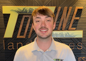 ndividual standing indoors in front of a wall with a large “TopLine Landscapes” logo, wearing a light-colored polo shirt with the company logo embroidered on the chest.