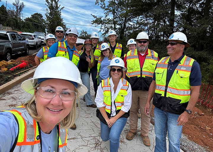 Safety Consultants USA team members wearing hard hats, safety glasses, and high-visibility vests gather at an active construction site during a field visit. The group stands on a newly constructed sidewalk with construction vehicles, utility work, and trees visible in the background.