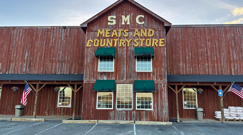 Front exterior of SMC Meats and Country Store, a large barn-style wooden building with weathered red siding, green window awnings, and American flags displayed near the entrance.
