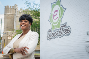 A person wearing a light-colored blazer stands outdoors beside a white brick wall featuring the EssenceTree logo. Trees and a large stone building are visible in the background.