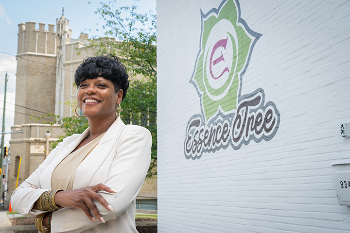 A person wearing a light-colored blazer stands outdoors beside a white brick wall featuring the EssenceTree logo. Trees and a large stone building are visible in the background.