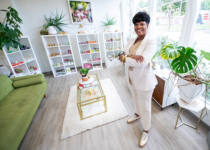 A person stands with arms crossed inside a retail showroom. White display shelves filled with skincare and wellness products line the walls, while indoor plants, a sofa, a coffee table, and large windows brighten the space.