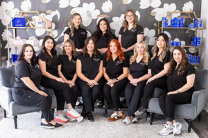 Southern Savvy Med Spa team members pose together in a spa reception area, wearing black branded uniforms and seated and standing in front of a floral-patterned wall with retail product displays.