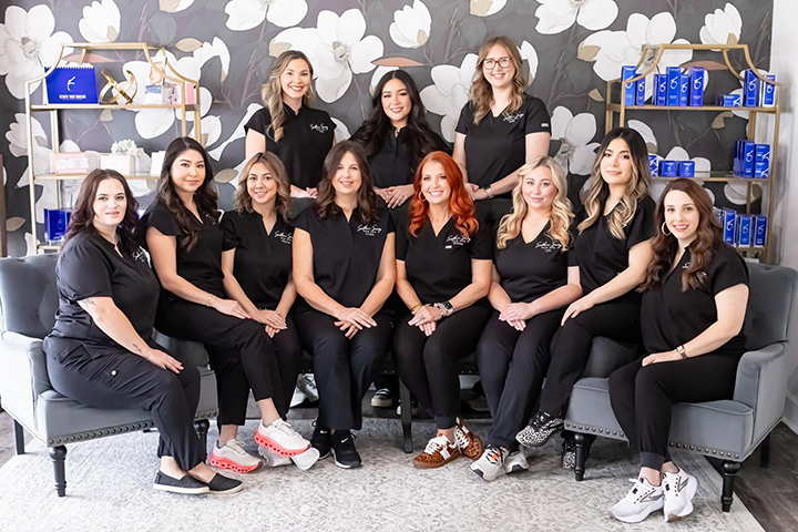 Southern Savvy Med Spa team members pose together in a spa reception area, wearing black branded uniforms and seated and standing in front of a floral-patterned wall with retail product displays.