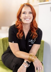 : Southern Savvy Med Spa co-owner Britney Exum sits in a green chair inside the spa, wearing a black branded uniform and jewelry, with a softly lit interior in the background.
