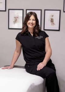 Southern Savvy Med Spa co-owner Kristie Parrish poses in a treatment room, seated on the edge of a spa treatment table with framed artwork displayed on the wall behind.
