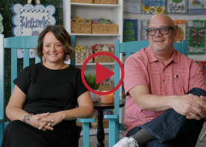 Drew and Kim Winters of DK Designs sit in turquoise rocking chairs inside their Forsyth, Georgia showroom. Decorative door hangers and home décor products designed by the company are displayed on shelves and walls in the background.